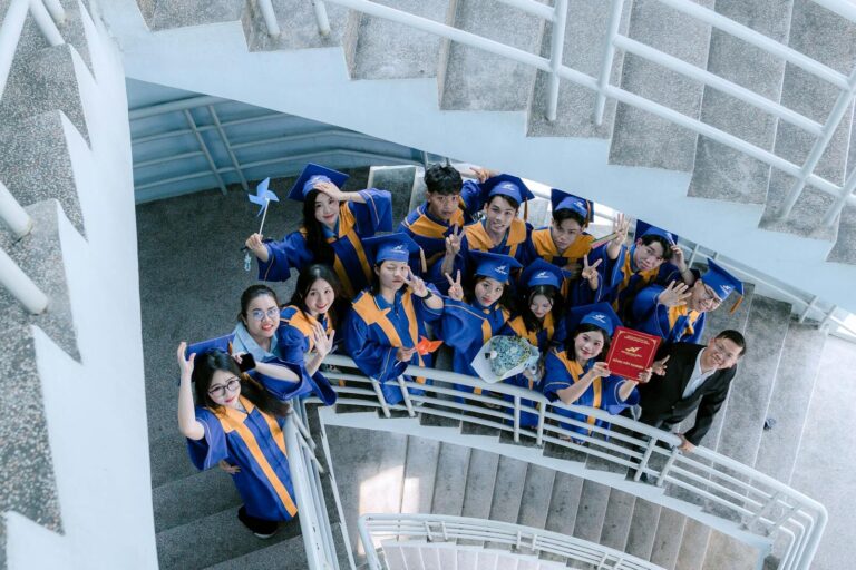 Cheerful graduates in blue gowns celebrate on a spiral staircase, capturing a joyous moment.